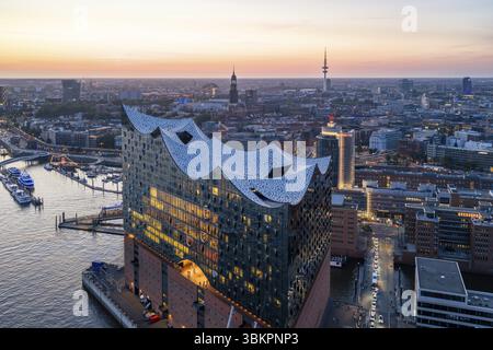 Luftaufnahme der Elbphilharmonie bei Sonnenuntergang mit beleuchteten Fenstern und Blick über Hamburg, Landungsbrücken und die Hauptkirche St. Michaelis (Miche Stockfoto