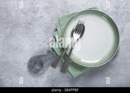 Ein leerer Teller und Besteck auf einem Schneidebrett, mit grüner Serviette, grauem Tisch, Blick von oben Stockfoto