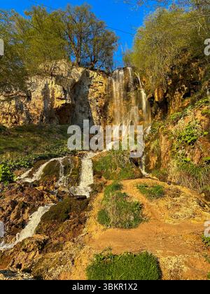 Ein malerischer Wasserfall, der über moosige Felsen stürzt, umgeben von üppigem Grün und Klippen unter einem klaren blauen Himmel, gefangen im warmen Sonnenlicht. Stockfoto
