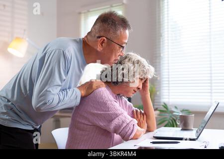 Ein liebevolles älteres asiatisches Paar unterstützt sich zuhause, während es ein Notebook benutzt. Ein fürsorglicher Partner hilft bei digitalen Aufgaben. Sie genießen das Leben im Ruhestand Stockfoto