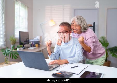 Ein älteres asiatisches Paar feiert einen Erfolg mit seinem Laptop im Home Office. Sie genießen ihren Ruhestand zusammen mit der modernen tec Stockfoto