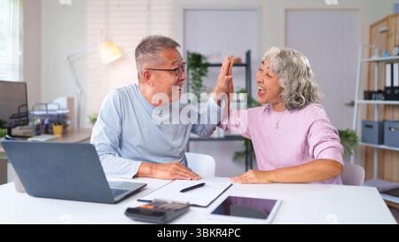 Fröhliches, älteres asiatisches Paar, das High Five feiert, um zu Hause finanziellen Erfolg zu feiern. Genießen Sie den Ruhestand, planen Sie Einsparungen, Investitionen zusammen mit h Stockfoto