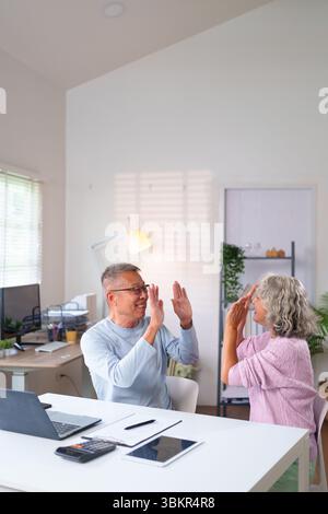Glückliches älteres asiatisches Paar feiert einen Sieg mit einer hohen fünf im Home Office und zeigt Freude. Sie planen ihren Ruhestand mit technischen Geräten. Stockfoto