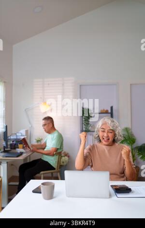 Eine ältere asiatische Frau feiert in ihrem Heimbüro Erfolg, während sie an einem Laptop arbeitet, während ein älterer Mann im Hintergrund ein Buch liest. Stockfoto