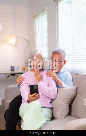 Fröhliches, älteres asiatisches Paar, das zu Hause ein Mobiltelefon für Videoanrufe benutzt. Ältere Frau, die ein Smartphone hält, lacht mit dem Mann im Wohnzimmer. Stockfoto