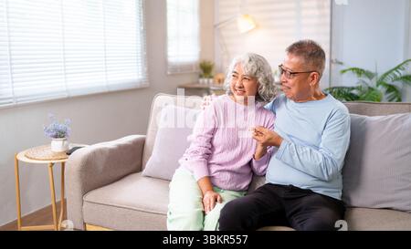 Ein älteres asiatisches Paar teilt sich einen liebevollen Moment auf dem Sofa im Wohnzimmer zu Hause. Sie sind glücklich, umarmen und genießen ihre gemeinsame Zeit in Komfort Stockfoto