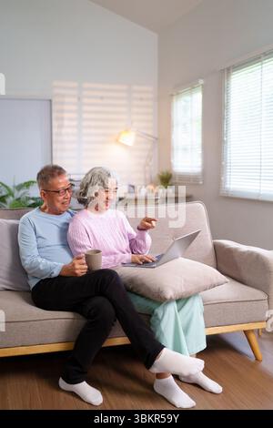 Älteres asiatisches Paar genießt eine gute Zeit zu Hause. Sie entspannen auf dem Sofa, benutzen einen Laptop, lächeln und trinken Kaffee. Happy Moments kreieren Last Stockfoto