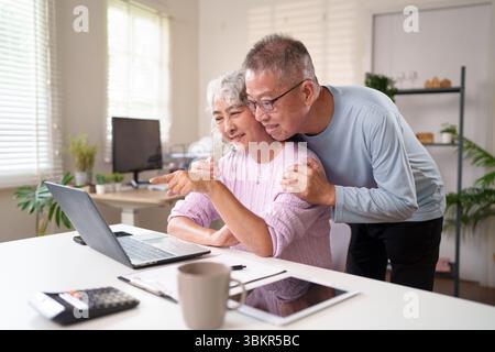 Ein asiatisches Seniorenpaar benutzt gern einen Laptop in ihrem Haus. Sie teilen einen zärtlichen Moment, während sie stöbern und lächeln und ihre gemeinsame Zeit genießen. Stockfoto