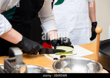 Nahaufnahme eines Küchenchefs mit schwarzen Handschuhen, der eine frische Aubergine auf einem Schneidebrett in einer professionellen Küche schneidet. Stockfoto