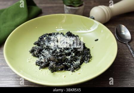 Risotto mit kräftiger schwarzer Tintenfischtinte und frisch geriebenem Parmesan, serviert in einer hellgrünen Schüssel auf einer hölzernen Essoberfläche. Stockfoto