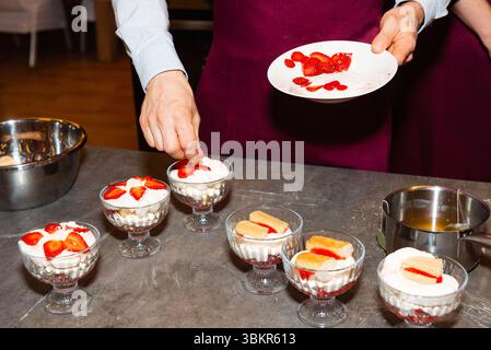 Nahaufnahme einer Person, die mehrschichtige Desserttassen mit frisch geschnittenen Erdbeeren und Sahne in einer Küchenumgebung garniert. Stockfoto