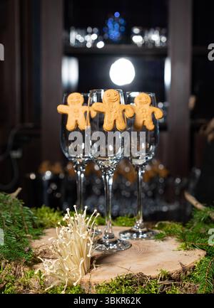 Vier leere Sektflöten, dekoriert mit Lebkuchenmännchen-Keksen, stehen auf einem festlichen Holzsockel mit Moos und Kiefern. Stockfoto