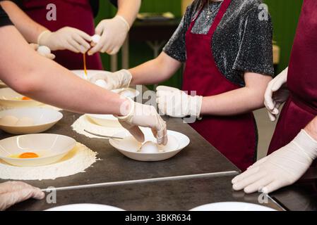 Die Teilnehmer eines Kochkurses mit Handschuhen und Schürzen bereiten Teig zu, indem sie Eier in Schüsseln knacken, die von Mehl umgeben sind. Stockfoto