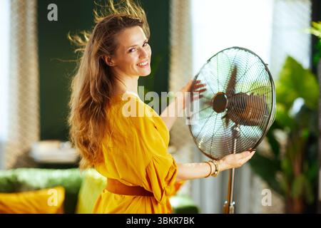 Eine lächelnde Frau mit langen braunen Haaren, die ein gelbes Kleid trägt, hält einen elektrischen Ventilator, ihr Haar weht, als sie erfrischenden Komfort und Glück findet Stockfoto