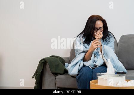 Eine kranke Frau mit Brille sitzt auf der Couch, niest in das Taschentuch, während sie ein Glas Wasser hält. In eine blaue Decke gehüllt, scheint sie müde zu sein. Konzept von h Stockfoto