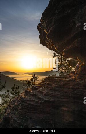 Sonnenaufgang über einer Berglandschaft. Aufgehende Sonne hinter einer Sandsteinklippe im Wald. Nebelbildung im Tal bei Rözenfelsen, Pfälzerwald, Stockfoto