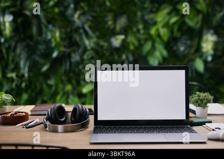 Nahaufnahme eines modernen Laptops mit leerem Bildschirm auf einem Holzschreibtisch, umgeben von kabellosen Kopfhörern, Topfpflanzen, Schreibwaren und Kaffeetasse im Büro mit grüner Wand, Kopierbereich Stockfoto