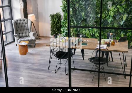 Moderner Business-Tagungsraum mit großem Holztisch mit digitalen Geräten, Notizbüchern, Kaffeetassen und grünen Pflanzen, leeren Stühlen um den Arbeitsbereich im modernen Büro Stockfoto