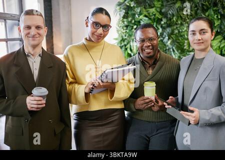 Porträt einer vielfältigen Gruppe junger Erwachsener und Profis mittleren Alters, die zusammen stehen und Kaffeetassen und ein digitales Tablet halten, in der modernen Büroumgebung in die Kamera lächeln Stockfoto