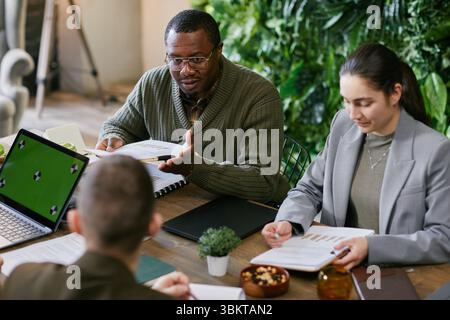 Gruppe von jungen Erwachsenen und multiethnischen Geschäftsleuten mittleren Alters, die während des Meetings über Finanzdokumente und Diagramme diskutieren, Schwarze Mann-Gesten, während Kollegen Notizen machen Stockfoto