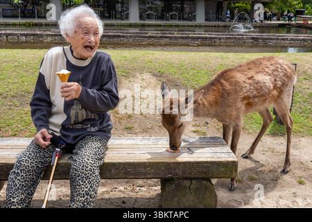 Eine alte japanerin isst Eis und lacht, während Sika-Hirsch im Nara Park in Japan einen Keks isst Stockfoto