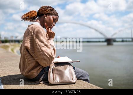 Die junge schwarze Frau sitzt am Fluss in der Stadt. Sie entspannt sich und macht Pläne. Stockfoto