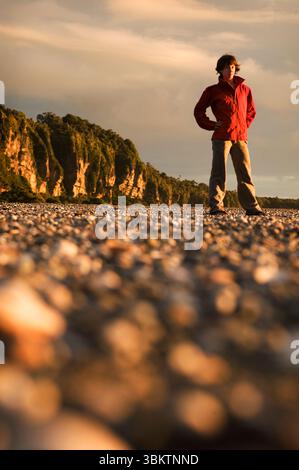 Eine junge Frau in roter Jacke steht an einem felsigen Strand bei Sonnenuntergang in Neuseeland. Stockfoto