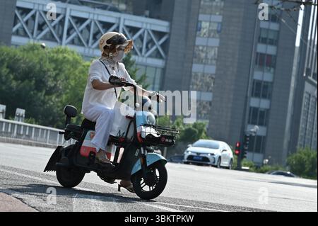 Peking, China. Juni 2025. Eine Frau mit einem Sonnenhut unter dem Helm und einer Maske auf dem Gesicht fährt mit einem Roller. Peking ist derzeit von extremer Hitze betroffen. Die Stadtverwaltung hat die Bewohner der chinesischen Hauptstadt gewarnt, dass die Temperaturen bis zu 38 Grad Celsius erreichen könnten. Quelle: Johannes Neudecker/dpa/Alamy Live News Stockfoto