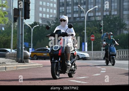 Peking, China. Juni 2025. Eine Frau in eine weiße Jacke gewickelt, mit Maske und Sonnenbrille fährt mit einem Roller. In China schützen sich viele Menschen mit langer Kleidung und Gesichtsmasken vor der Sonne - auch um einen hellen Hautton zu erhalten, der in China beliebt ist. Peking ist derzeit von extremer Hitze betroffen. Die Stadtverwaltung warnte die Bewohner der chinesischen Hauptstadt vor Hochtemperaturen von bis zu 38 Grad. In der Folge wurde der Sportunterricht beispielsweise in Schulen und Kindergärten abgesagt. Quelle: Johannes Neudecker/dpa/Alamy Live News Stockfoto