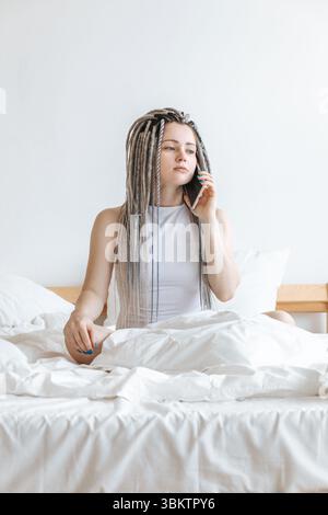 Junge Frau, die am Telefon im weißen Bett spricht. Stockfoto