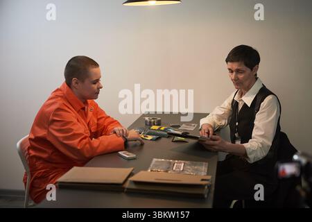Kaukasischer junger erwachsener Mann in orangefarbener Gefängnisuniform, sitzt am Tisch mit einer kaukasischen Offizierin mittleren Alters, die während der Verhörszene Dokumente überprüft Stockfoto