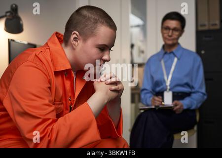 Junge kaukasische Frau, die in orangefarbener Gefängnisuniform sitzt, mit gefalteten Händen, nachdenklich erscheint, während Kaukasierin mittleren Alters in Brille im Hintergrund beobachtet und Notizen macht Stockfoto