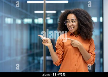 Eine glückliche Frau zeigt zur Seite, lächelt mit einer Brille in einem Büro. Stockfoto