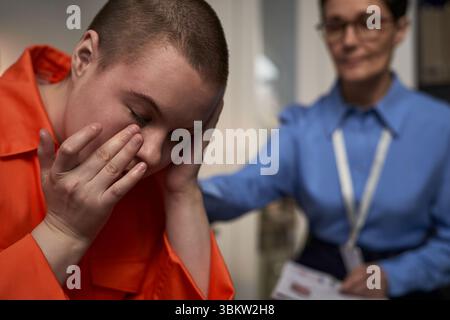 Kaukasische junge Erwachsene Frau in Gefängnisuniform sitzend mit dem Kopf in den Händen, während kaukasische Frau mittleren Alters in Uniform in der Nähe steht und Unterstützung anbietet Stockfoto