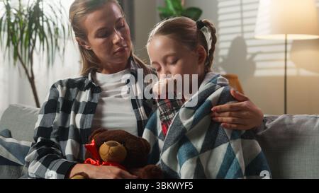Kaukasische Familie kuschelt zu Hause fürsorgliche Mutter tröstet kranke Tochter Husten krank eingewickelt in Decke auf Couch trauriges Kind Kind Mädchen umarmt Teddy Stockfoto
