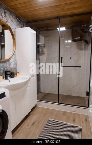 Das helle, moderne Badezimmer verfügt über eine rahmenlose Glasdusche mit Marmorfliesen, einen weißen Kosmetikbereich mit Waschbecken und ein warmes Holzboden Stockfoto
