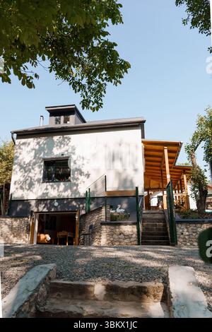 Stilvolles zweistöckiges Öko-Haus mit grauem Äußeren, Steinfundament, Zugang zur Holzterrasse über Treppen, üppiger Gartenumgebung und klarem blauem Himmel, der so Werbung macht Stockfoto