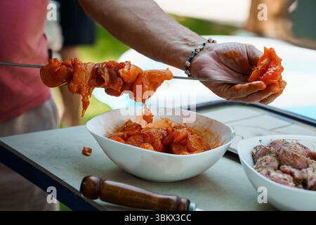 Ein Mann bereitet Hähnchenspieße zu, indem er würfeltes Fleisch mit einer leuchtenden roten Marinade überreicht, um im Sommer im Freien in einer Gartenumgebung zu grillen Stockfoto