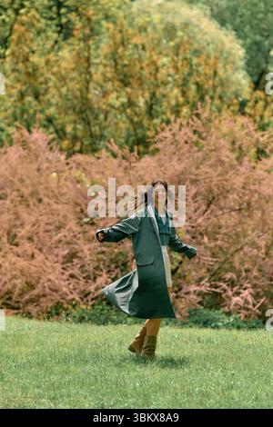 Eine junge Frau mit lockigen Haaren wirbelt fröhlich in einem stilvollen Regenmantel an einem sonnigen Tag im Freien. Stockfoto