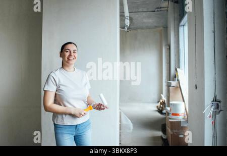 Eine Frau mittleren Alters hält eine Malerwalze und lächelt. Eine junge Dame steht in einem hellen, teilweise renovierten Zimmer mit unvollendeten Wänden und Fenstern. Buil Stockfoto