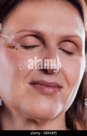 Eine junge Frau trägt das Serum auf ihr Gesicht auf. Eine Frau mittleren Alters hält einen Tropfer mit einer feuchtigkeitsspendenden Essenz. Die Dame hat die Augen geschlossen und ist enjo Stockfoto