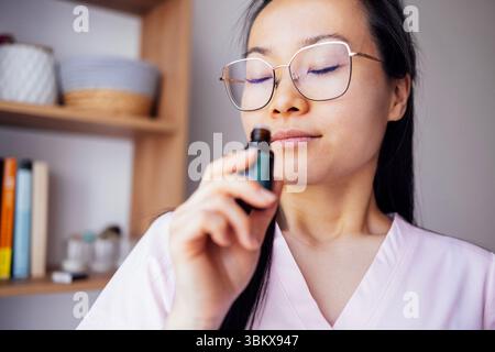 Eine junge asiatische Frau in lässiger Kleidung, die nach ätherischem Öl riecht. Eine attraktive koreanische Frau mit Brille schließt die Augen und atmet den Duft ein Stockfoto