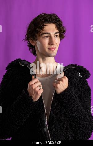 Studioaufnahme in Hüfthöhe eines jungen männlichen Erwachsenen, der eine schwarze flauschige Jacke trägt und von der Kamera wegblickt. Er hat dunkle lockige Haare und hält die Kante Stockfoto