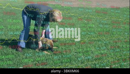 Eine sich beugende ältere Frau, die einen kleinen braunen Welpen auf einem breiten Grasfeld mit einem Welpenkragen hält Stockfoto