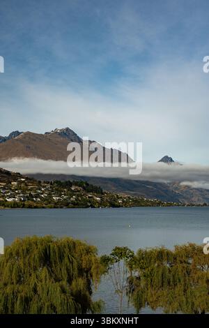 Ein ruhiger See mit Trauerweiden im Vordergrund und eine Stadt am Hügel unterhalb der hohen Berge, bedeckt von einer auffälligen Schicht niedriger Wolken unter einem Stockfoto