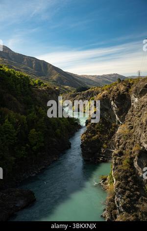 Ein pulsierender türkisfarbener Fluss schlängelt sich durch einen steilen Canyon, der von üppigen grünen Bäumen und zerklüfteten braunen Hügeln gesäumt ist, unter einem klaren blauen Himmel, der das Drama verdeutlicht Stockfoto