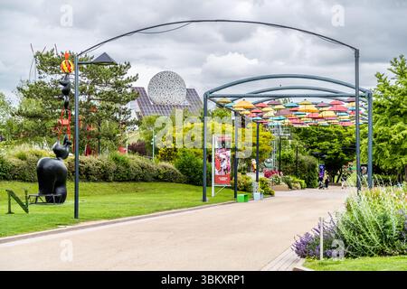 Blick auf einen Parkweg am Futuroscope (Chasseneuil-du-Poitou, Nouvelle-Aquitaine, Frankreich): Im Hintergrund befindet sich die markante gezähnte geodätische Kuppel des Parks, links eine abstrakte Skulptur und rechts eine Pergola mit bunten Sonnenschirmen. Die Kuppel gilt als Wahrzeichen des 1987 eröffneten Freizeitparks. Das Futuroscope bei Poitiers ist ein Vergnügungspark, der sich auf futuristische Themen, Multimedia-Erlebnisse und innovative Attraktionen spezialisiert hat Stockfoto