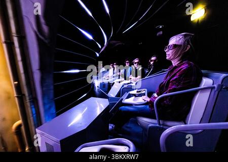 Besucher, die eine 3D-Brille in einem beweglichen Fahrgestell tragen, auf einer Simulatorfahrt im Freizeitpark Futuroscope. Blaue und violette Lichtstreifen entstehen durch die schnelle Bewegung. Die Fahrgeschäfte des Parks verbinden kinetische Plattformen mit einer großformatigen Stereoprojektion. Das Futuroscope bei Poitiers ist ein Vergnügungspark, der sich auf futuristische Themen, Multimedia-Erlebnisse und innovative Attraktionen spezialisiert hat. Avenue René Monory, Chasseneuil-du-Poitou, Vienne, Nouvelle-Aquitaine, Frankreich Stockfoto