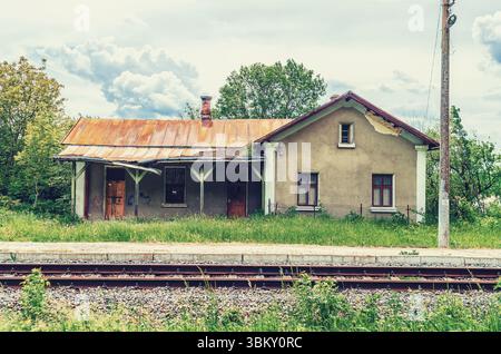 Ländlicher Blick auf den verlassenen Bahnhof mit Ziegelbau, mit Zinn bedeckt, mit kaputten Fenstern unter wolkenblauem Himmel und Eisenbahngleise Stockfoto