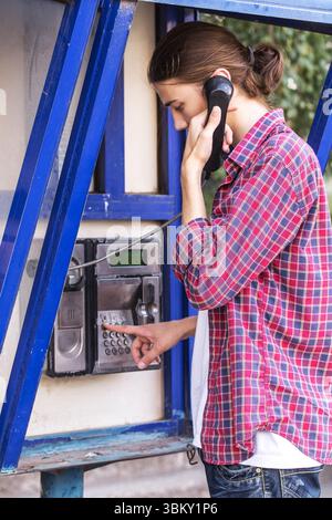 Junger gutaussehender bärtiger Mann in rotem Karohemd, der auf einem öffentlichen Telefon die Nummer wählt. Stockfoto